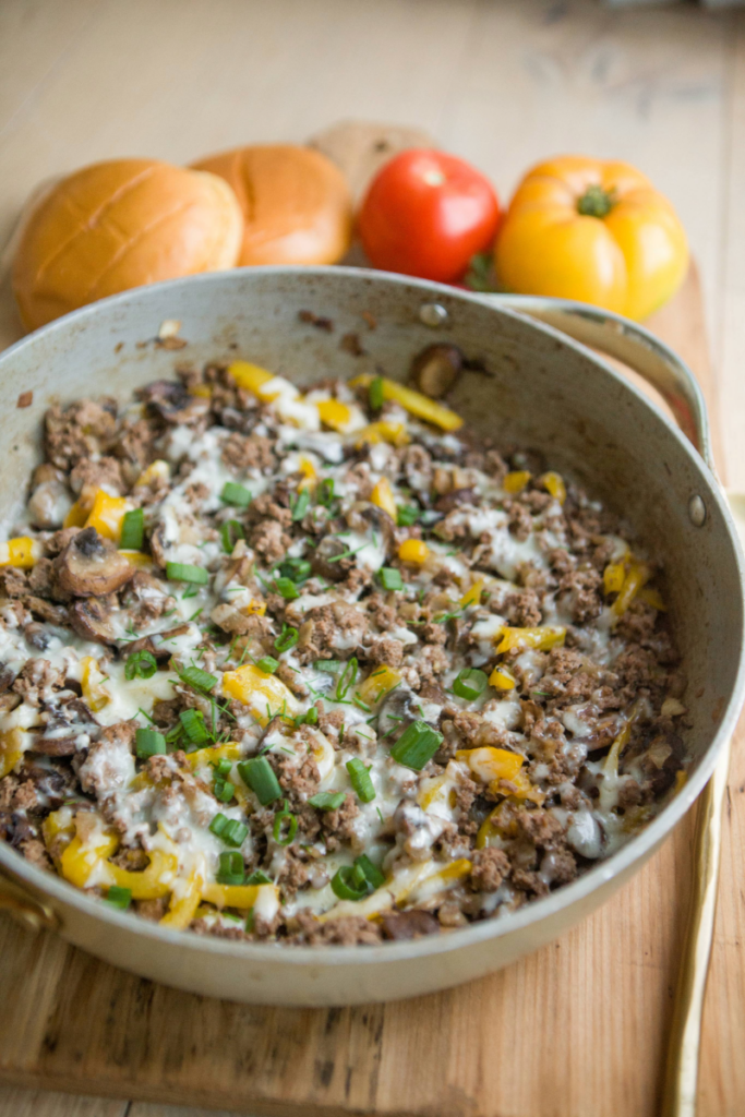Pan of philly ground beef skillet on top of wooden cutting board with buns and tomatoes off to the side.