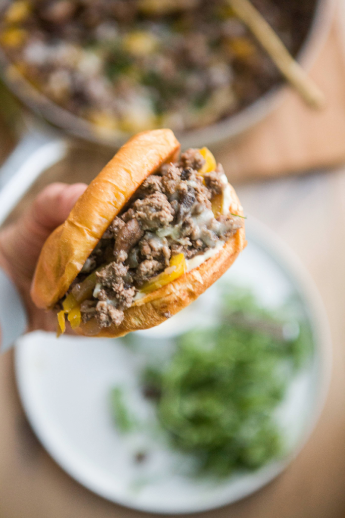 Philly ground beef skillet on brioche bun being held up close to the photo with plate with green in the background.