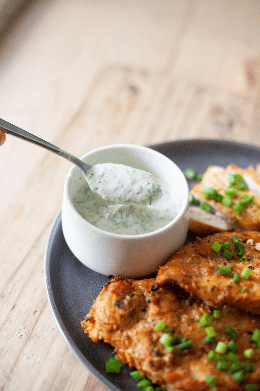 Herb yogurt dressing in a bowl on a plate next to cooked chicken.