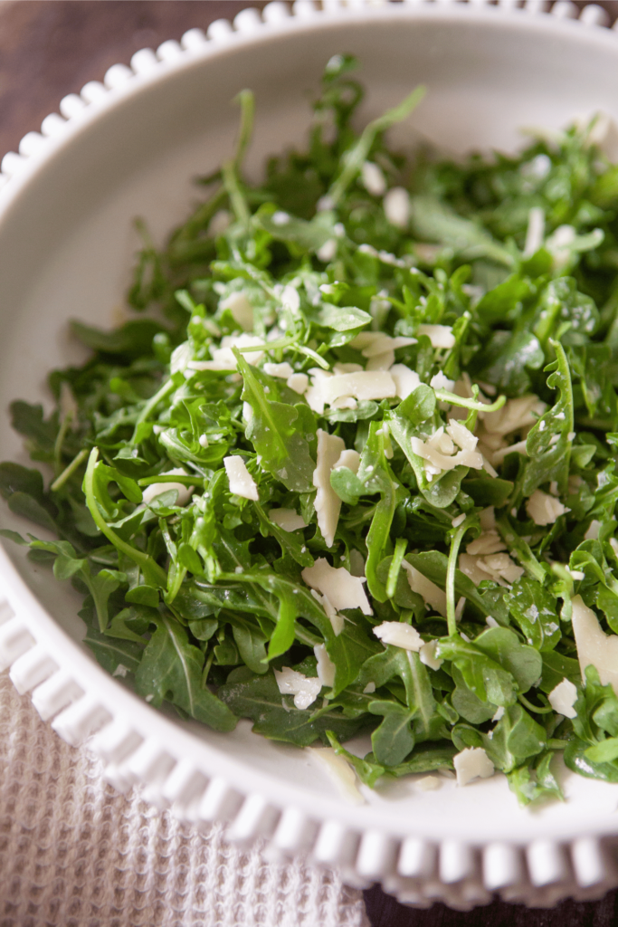 Close up of arugula salad topped with shaved parmesan cheese in a white bowl.