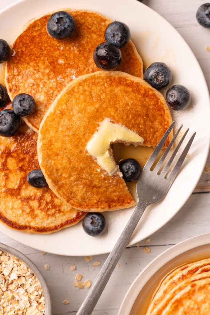 Plate of cottage cheese pancakes with butter and blueberries on plate with fork and bite taken out.