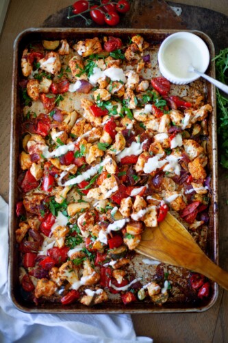 Sheet pan of harissa chicken drizzled with whipped feta hummus sauce with wooden spoon and bowl on hummus sauce and spoon in the corner of the pan.
