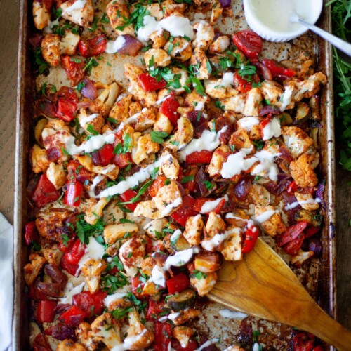 Sheet pan of harissa chicken drizzled with whipped feta hummus sauce with wooden spoon and bowl on hummus sauce and spoon in the corner of the pan.