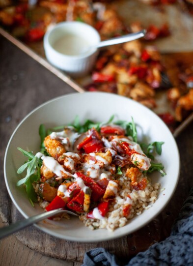 Harissa chicken and vegetables in white bowl with rice and arugula drizzled with whipped feta hummus sauce with fork. Sheet pan of chicken and hummus sauce behind white bowl.