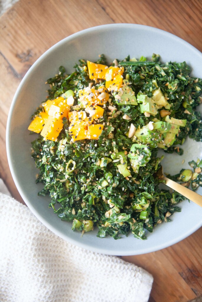 Kale salad topped with mango, peanuts and avocado in white bowl with gold spoon.