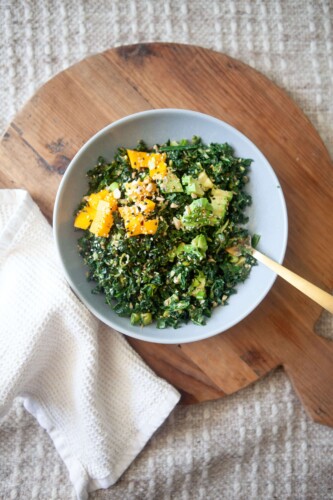 Kale salad with peanut vinaigrette in white bowl on wood cutting board with gold fork and white napkin.