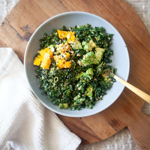 Kale salad with peanut vinaigrette in white bowl on wood cutting board with gold fork and white napkin.