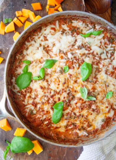 Butternut lasagna in metal skillet garnished with fresh basil on wooden board with cubes of butternut squash on board.