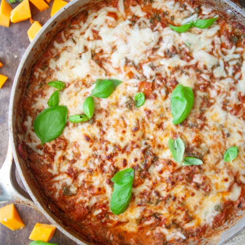 Butternut lasagna in metal skillet garnished with fresh basil on wooden board with cubes of butternut squash on board.