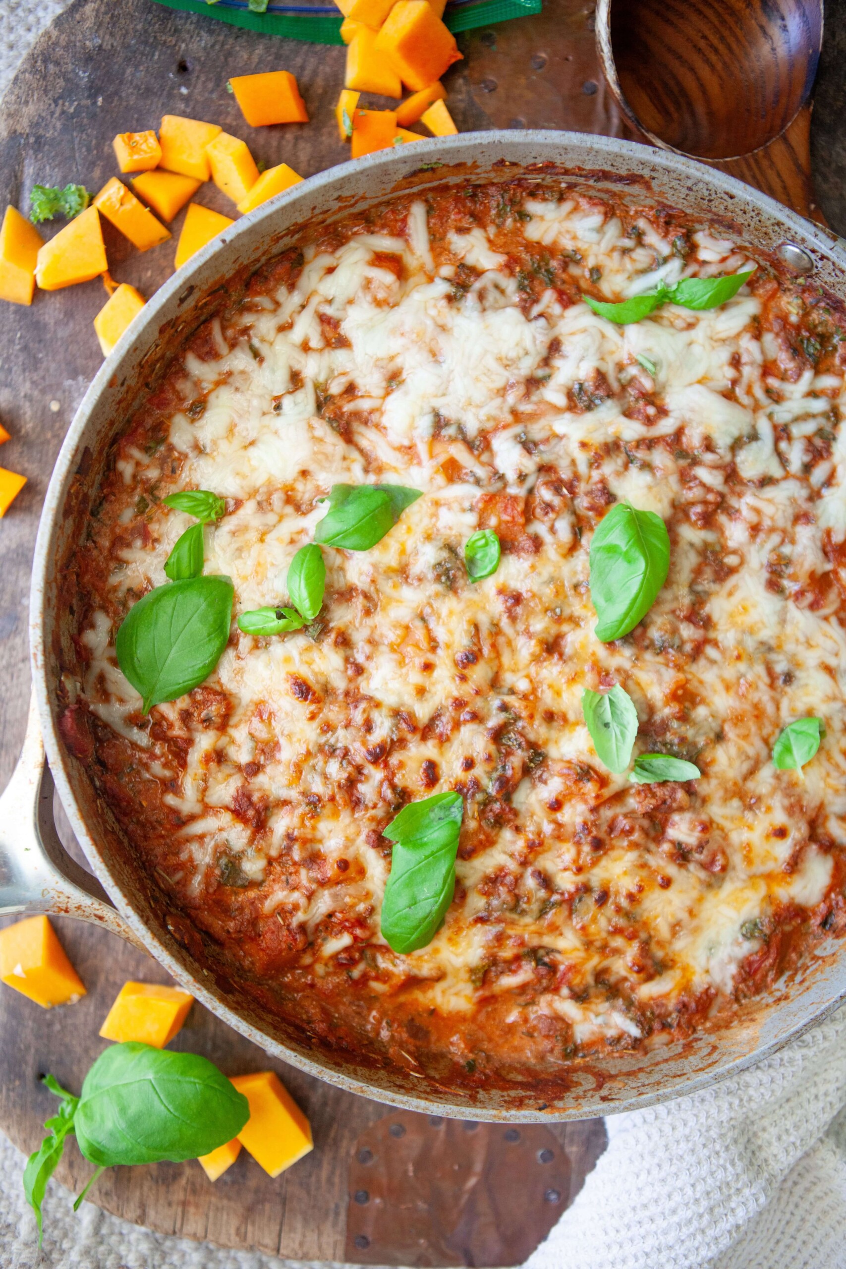 Butternut lasagna in metal skillet garnished with fresh basil on wooden board with cubes of butternut squash on board.