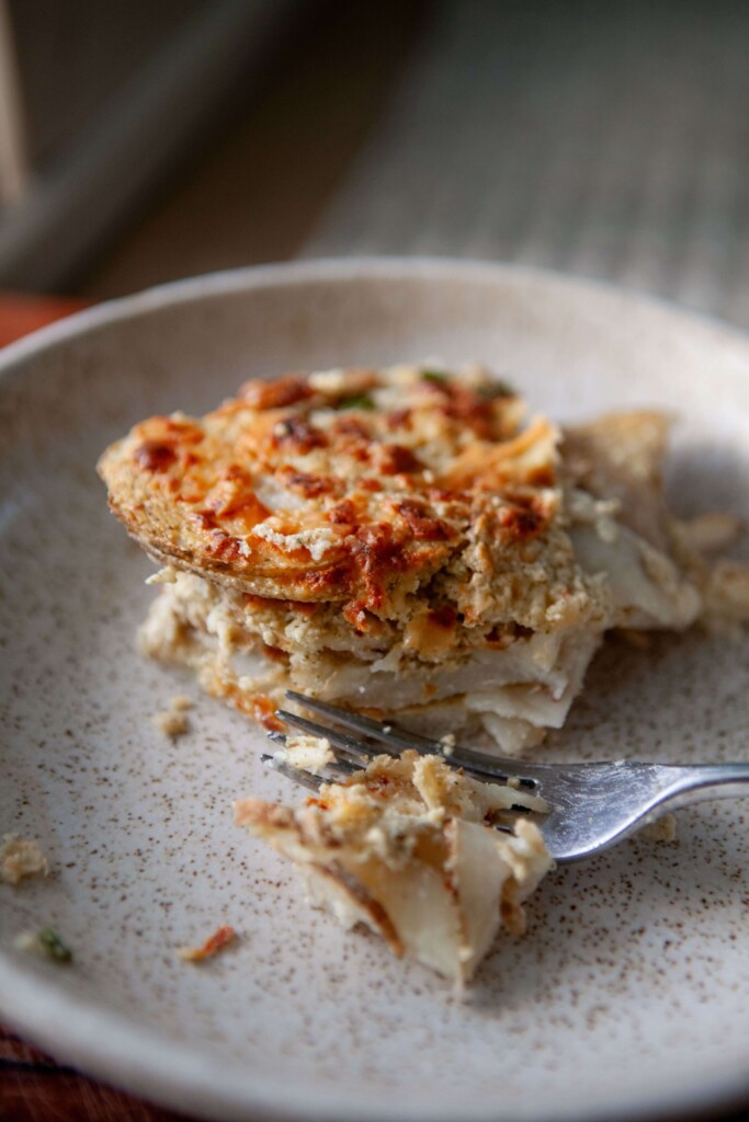 Fork cutting scalloped potatoes on ceramic plate.