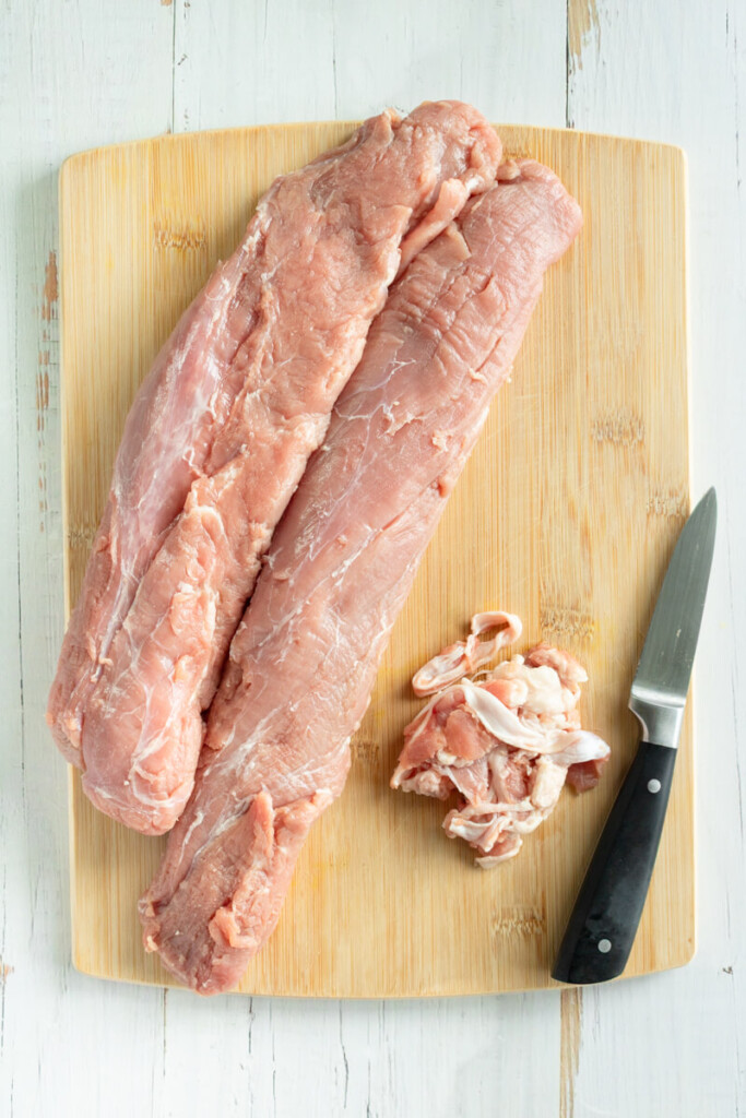Trimming excess fat off pork tenderloins on cutting board with knife.