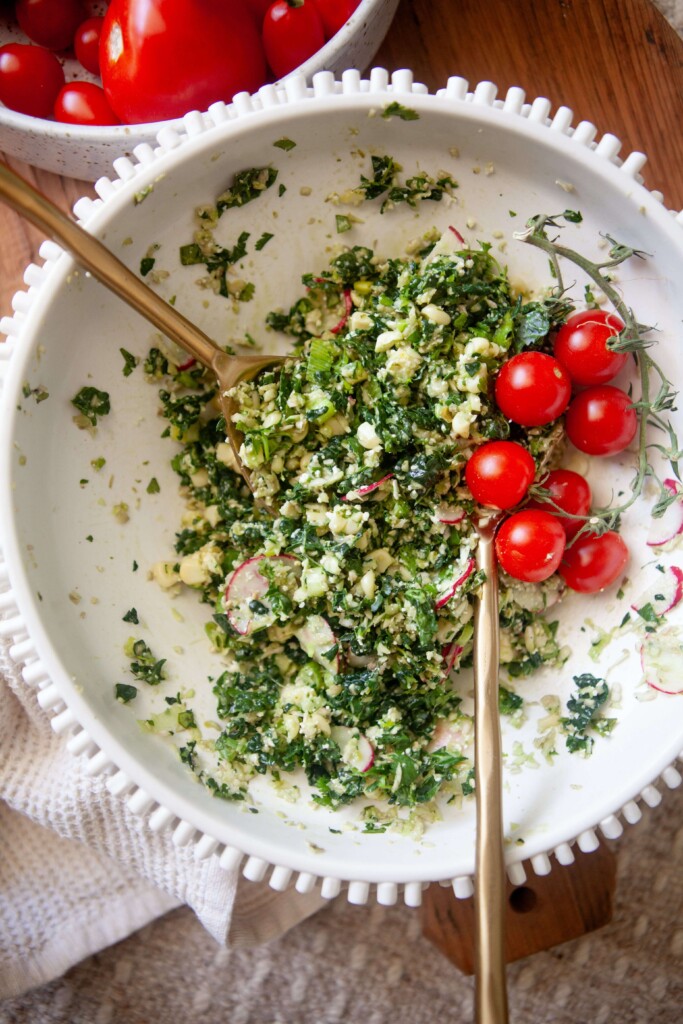Mexican chopped kale salad mixed together with two gold spoons in white ceramic bowl garnished with red cherry tomatoes.