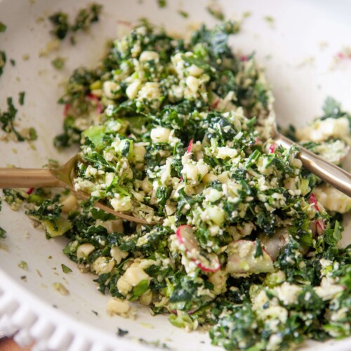 Mexican kale salad mixed together with two gold spoons in white ceramic bowl.
