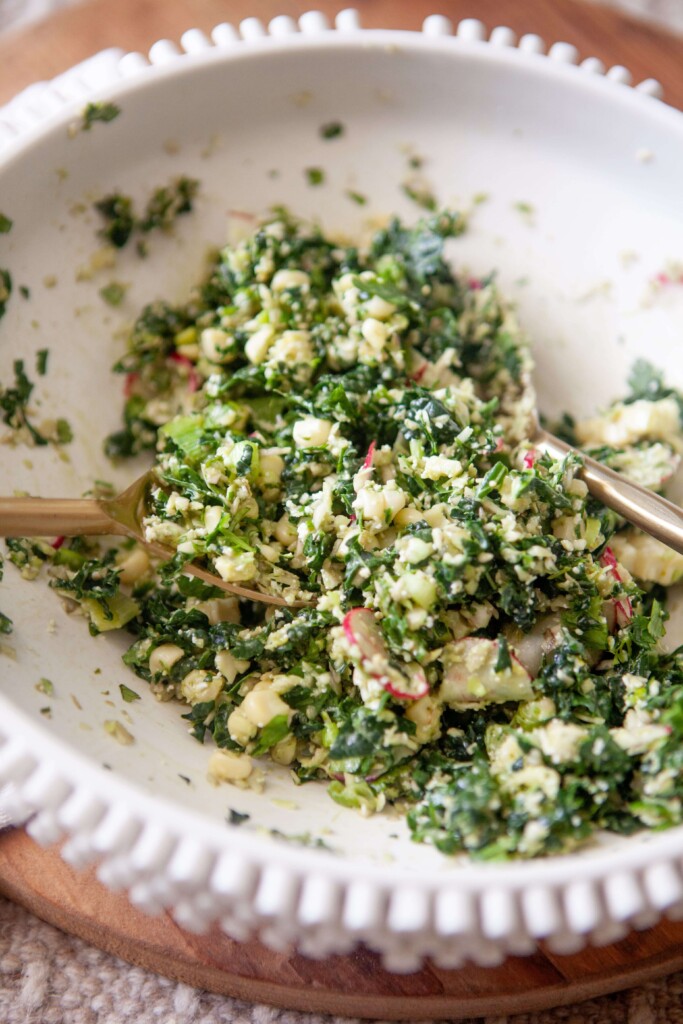 Mexican kale salad mixed together with two gold spoons in white ceramic bowl.