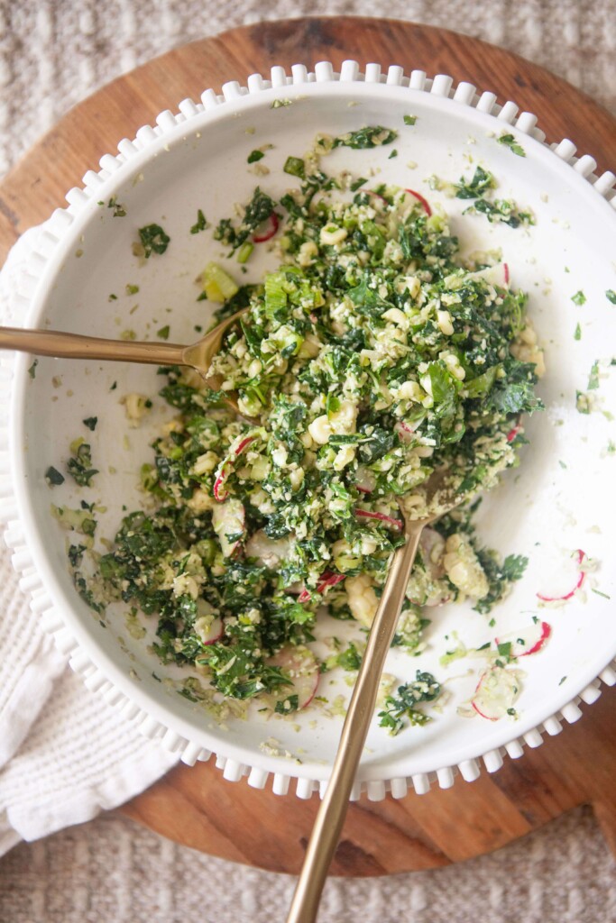 Mexican chopped kale salad mixed together with two gold spoons in white ceramic bowl.