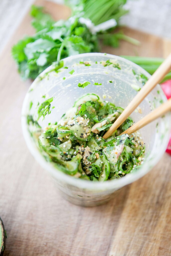 Pair of chopsticks in a plastic container of chicken avocado cucumber salad sprinkled with everything but the bagel seasoning on a wood board.
