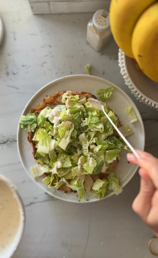 Adding additional greek yogurt caesar dressing to caesar salad ground chicken pizza on white plate.