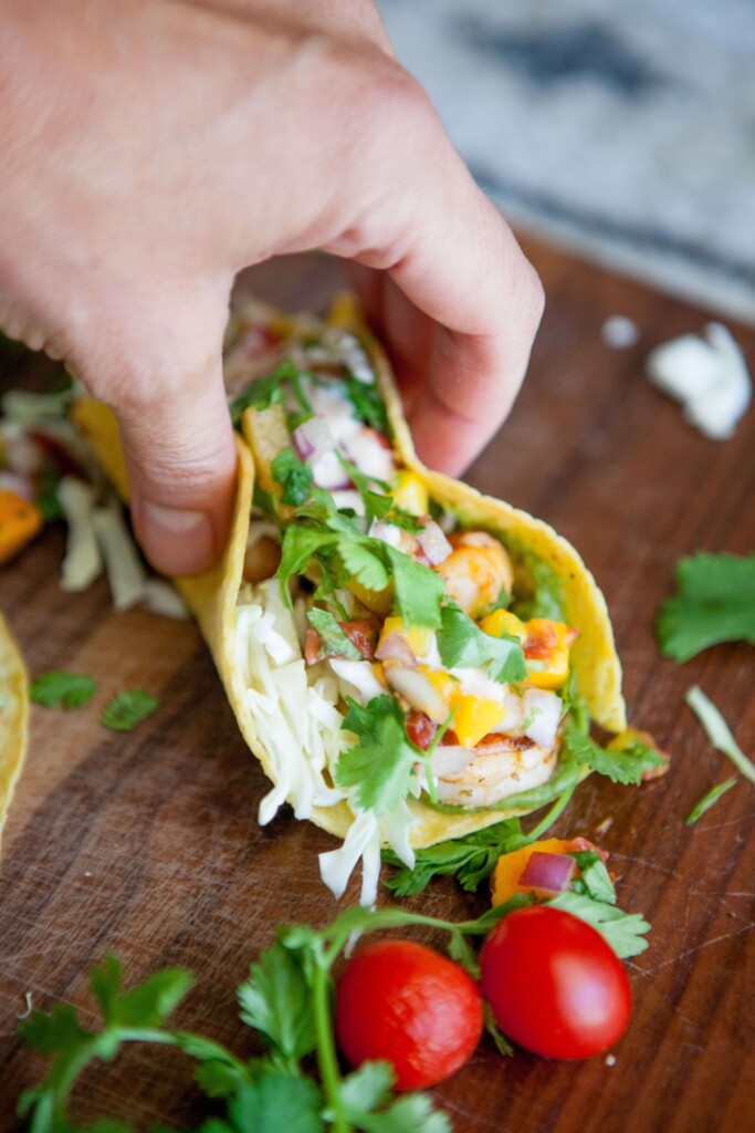 Hand holding shrimp taco on wood board.