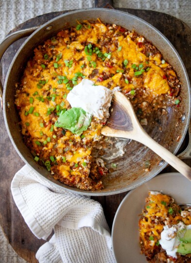 Skillet mexican potatoes in large skillet topped with sour cream and avocado with wood spoon and plate beside skillet with small serving.