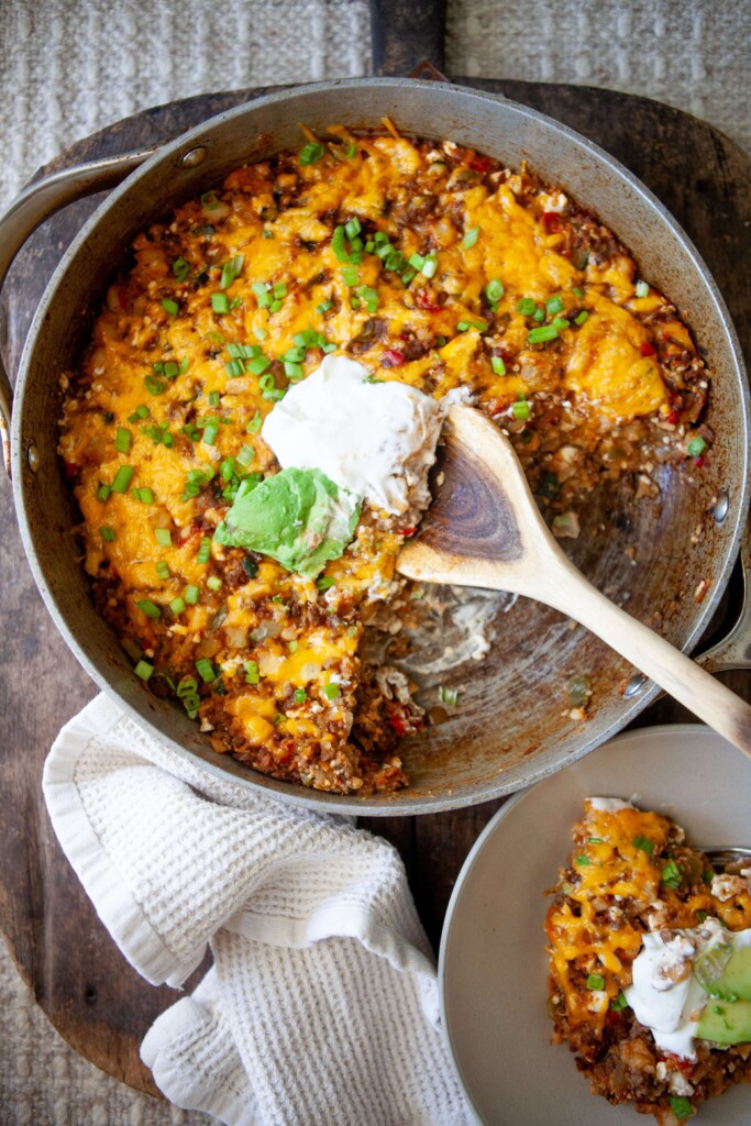 Skillet mexican potatoes in large skillet topped with sour cream and avocado with wood spoon and plate beside skillet with small serving.