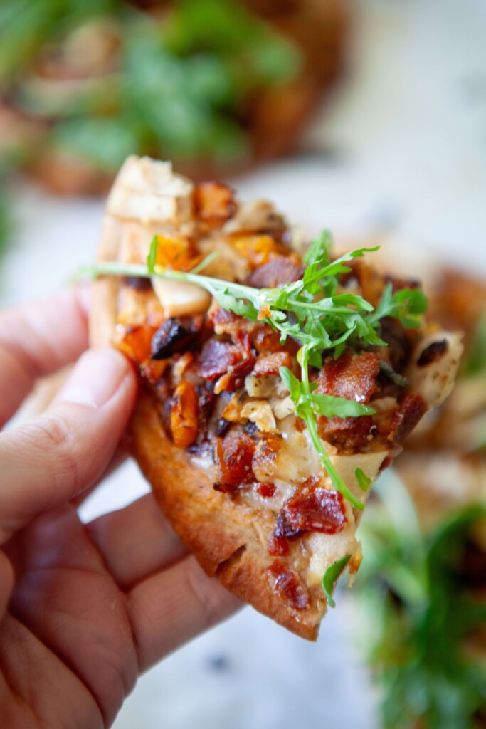 Hand holding a piece of a butternut squash, bacon, and caramelized onion flatbread.