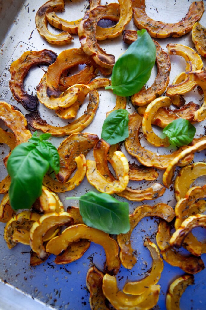 Roasted delicata squash half moons with caramelized edges on a metal sheet pan, sprinkled with dried herbs and flaky salt and topped with fresh basil leaves.