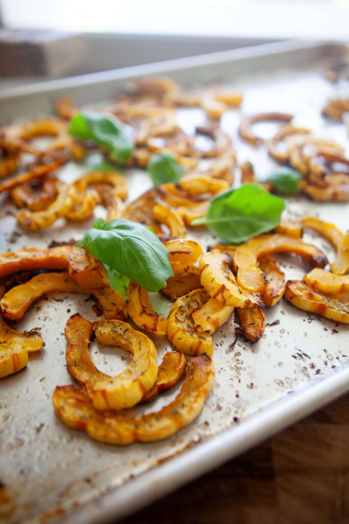 Close-up of roasted delicata squash slices on a sheet pan, showing the striped edible skin, golden caramelized edges, and fresh basil leaves.