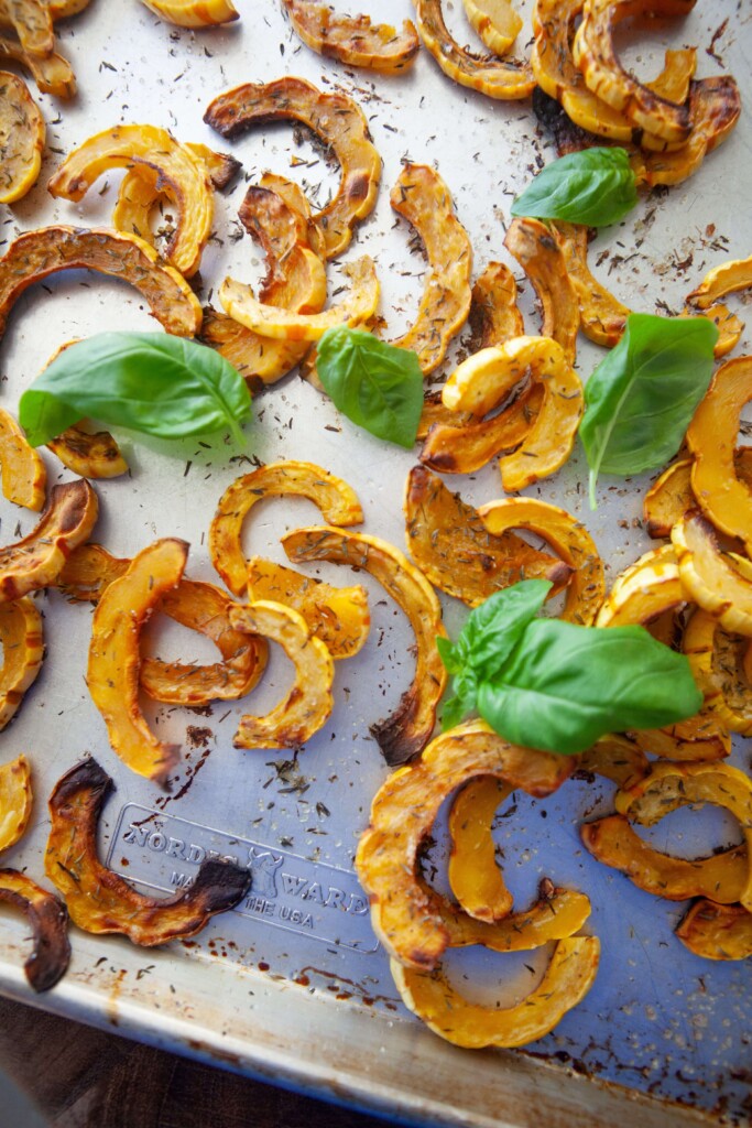Overhead view of roasted delicata squash slices spread in a single layer on a baking sheet, with browned edges, dried herbs, and fresh basil.