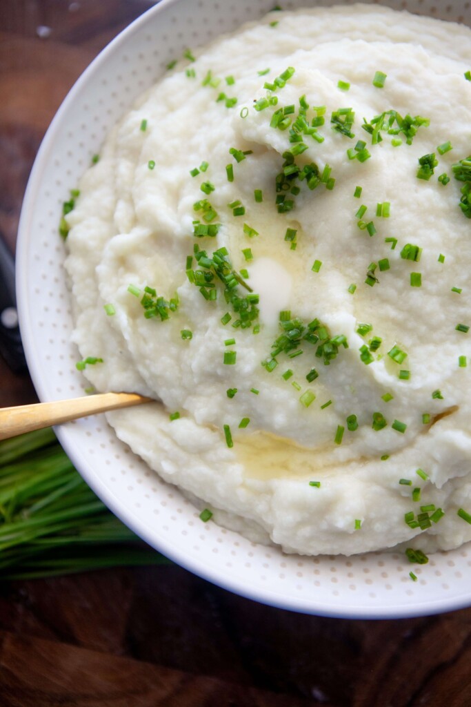 Mashed potatoes and cauliflower in white bowl with gold spoon.