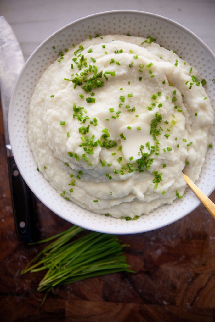 Mashed potatoes and cauliflower garnished with fresh chives in serving bowl with gold spoon.