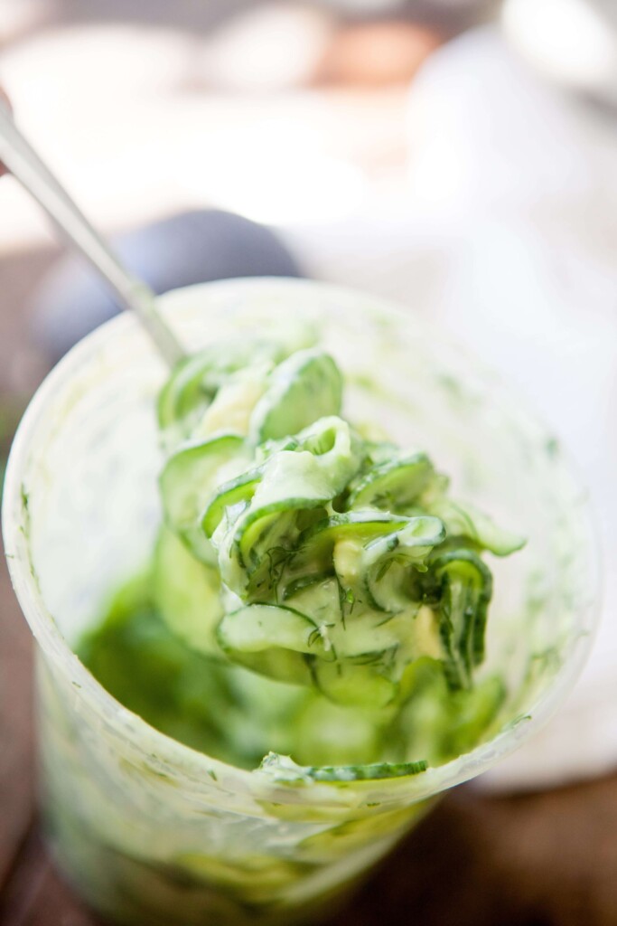 Spoon holding up creamy cucumber salad out of large deli container.