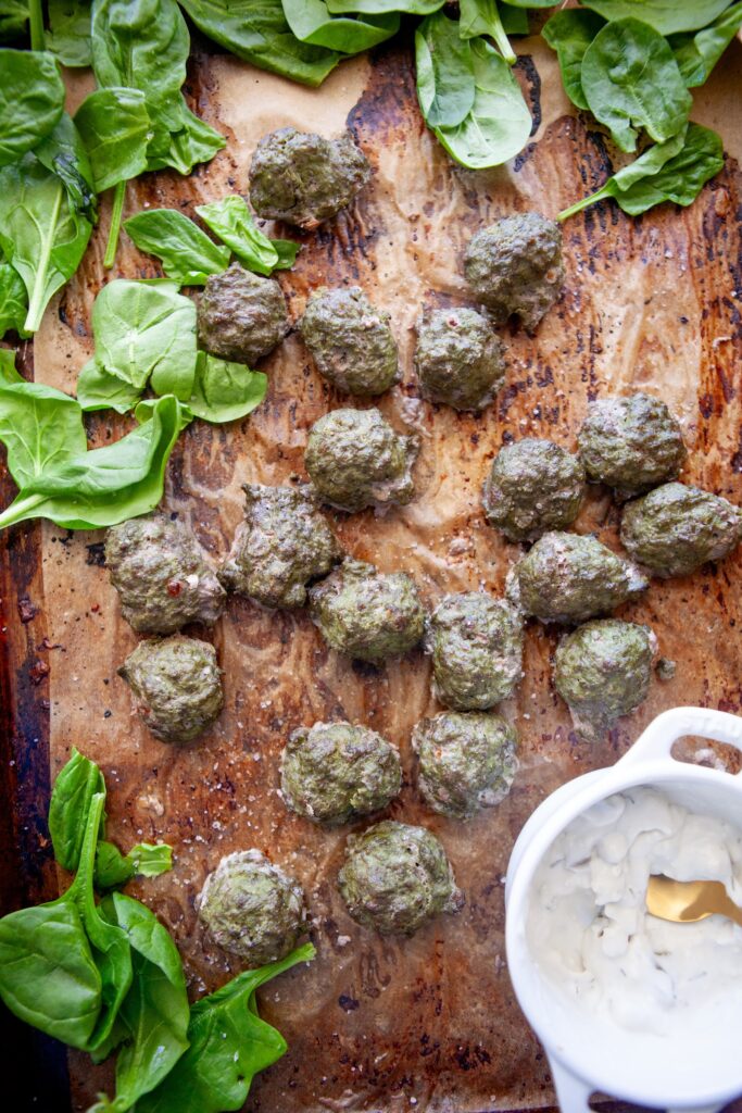 Overhead view of spinach basil meatballs on a parchment-lined baking sheet with fresh spinach and a bowl of yogurt sauce