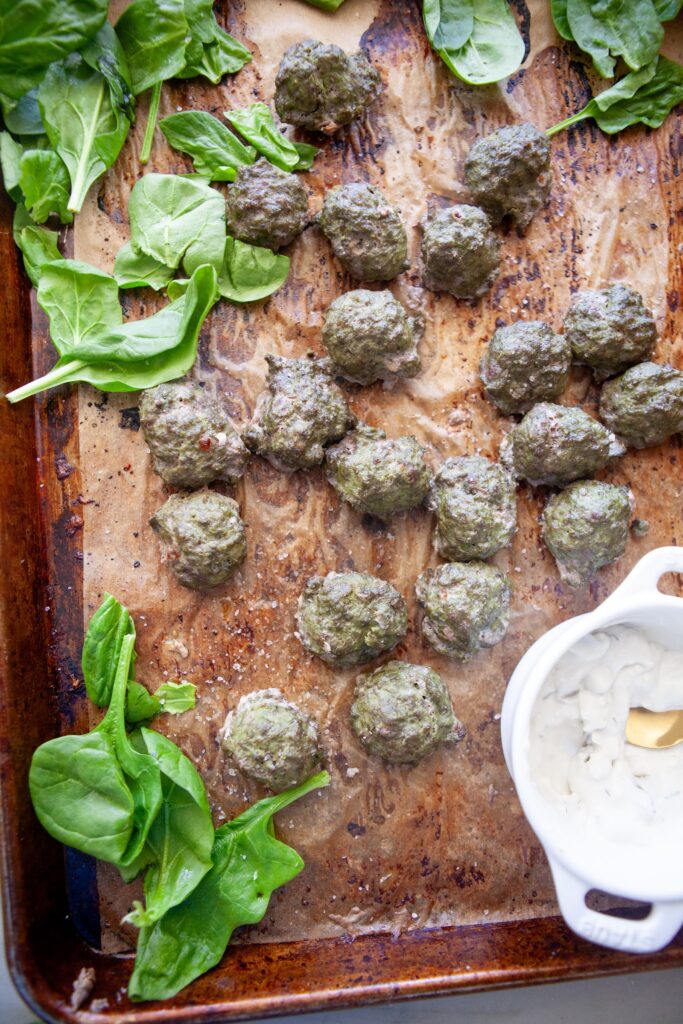 Sheet pan of high-protein spinach basil meatballs with spinach leaves and yogurt dip