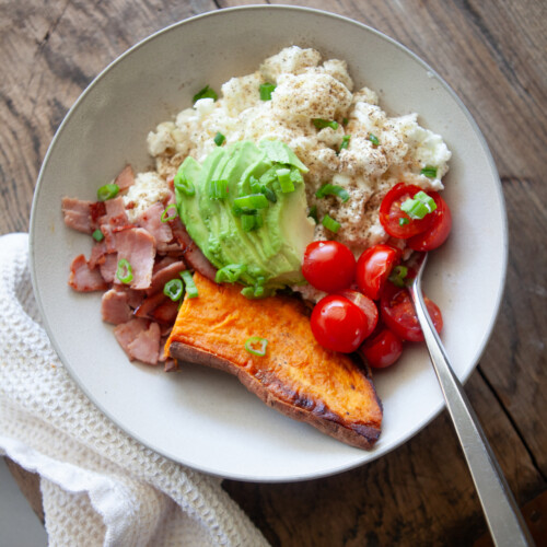 A high protein breakfast bowl featuring scrambled egg whites, chopped ham, avocado, cherry tomatoes, and a baked potato wedge topped with green onions, presented in the style of a hearty broccoli cheese baked potato meal.