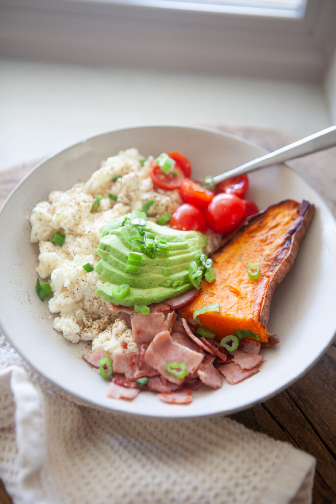 A high protein breakfast bowl containing scrambled egg whites, chopped ham, sliced avocado, cherry tomatoes, and a roasted sweet potato wedge garnished with green onions.